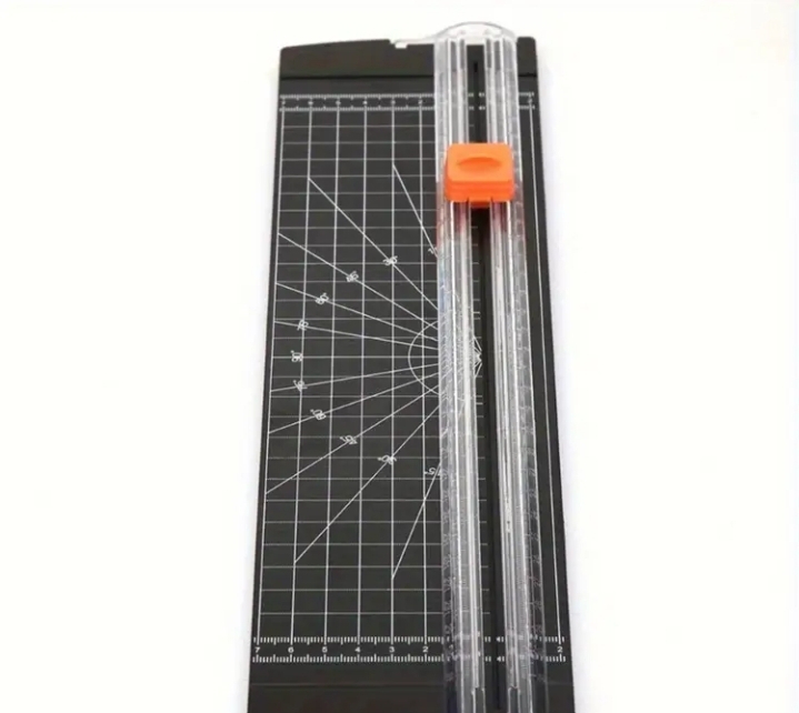 A paper trimmer featuring a measurement grid and a sliding blade with an orange handle.
